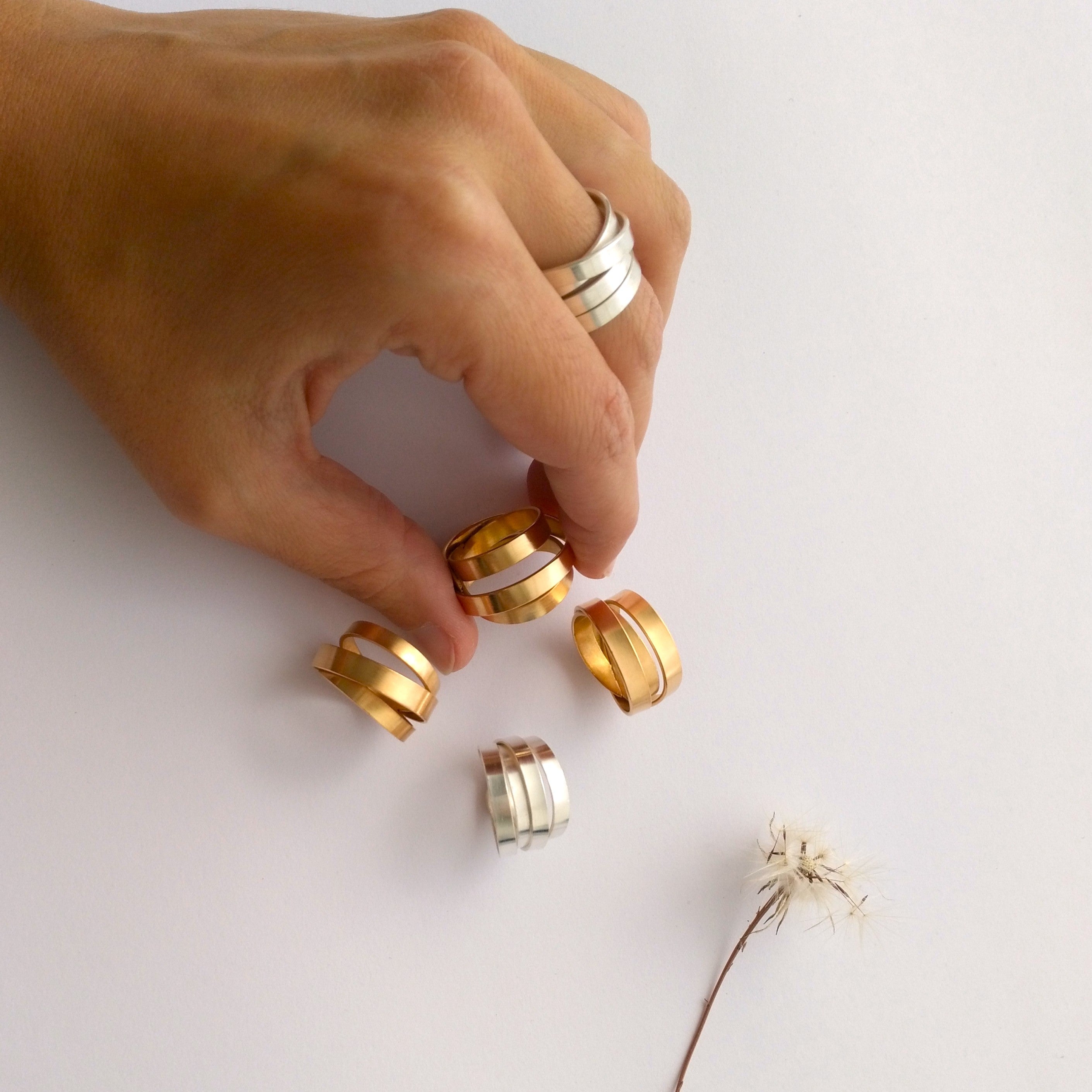 Hand holding mixed metal rings arranged on a light surface beside a small dried sprig.